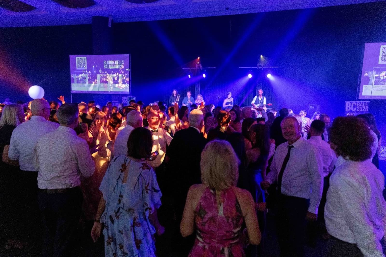 Crowd dancing in front of the band at the 2025 Bloody Cells Ball with the BC Ball lockup on the screen behind