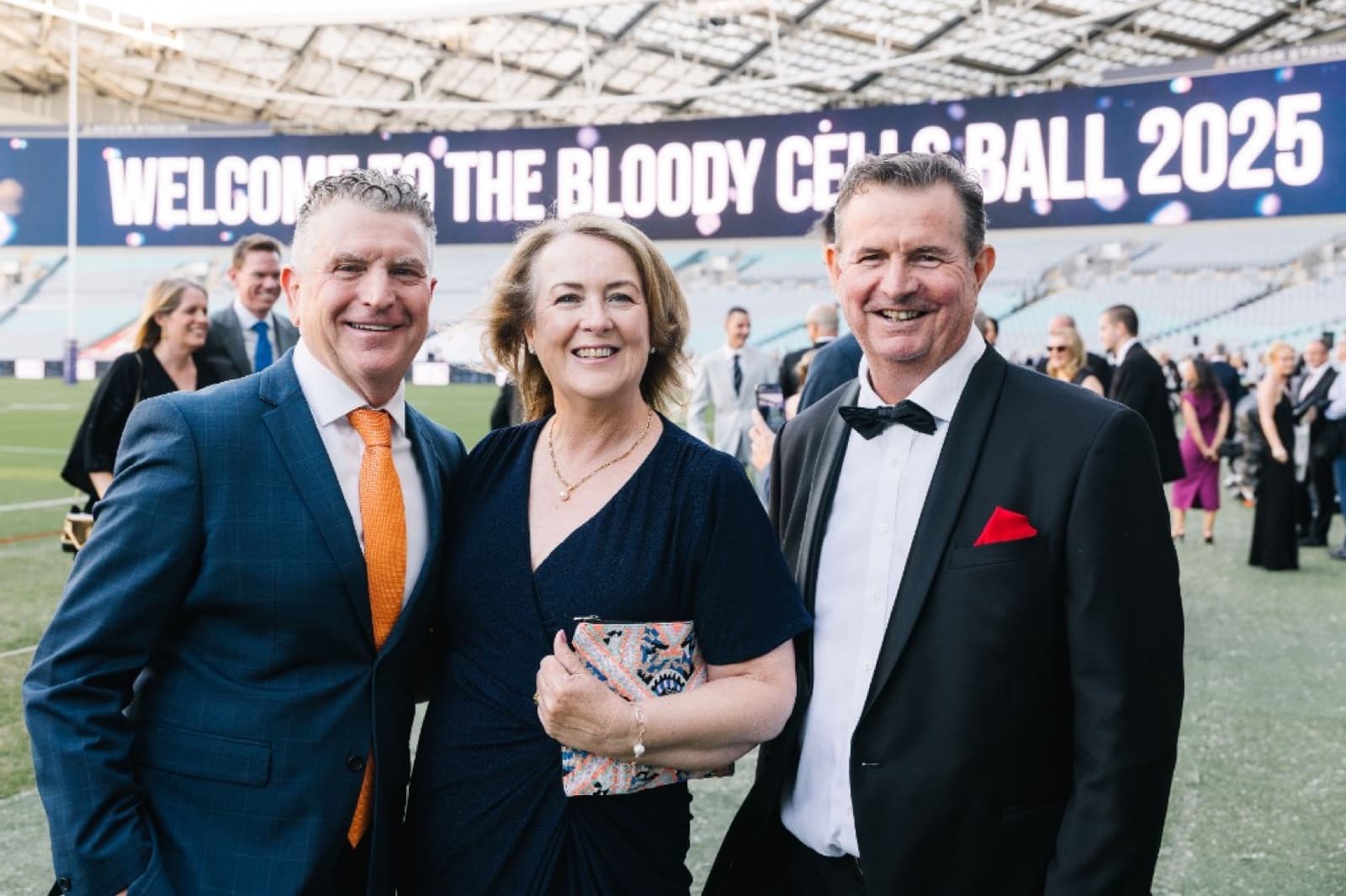 Three guests photographed in front of the 'Welcome to the Bloody Cells Ball 2025' Accor Stadium media wall