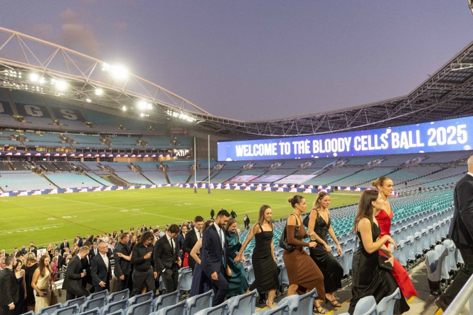 Guests walking down through Accor Stadium toward the dinner room as the 'Welcome to the Bloody Cells Ball 2025' screen plays overhead