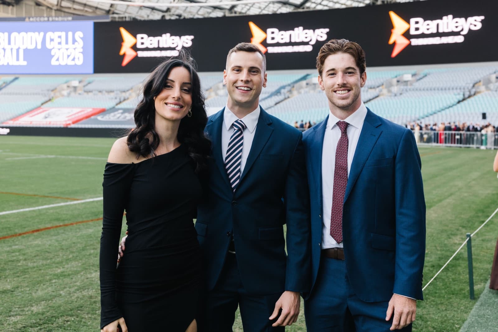 Three sponsor guests photographed on the Accor Stadium field with the Bentleys sponsor branding on the stadium screens behind them