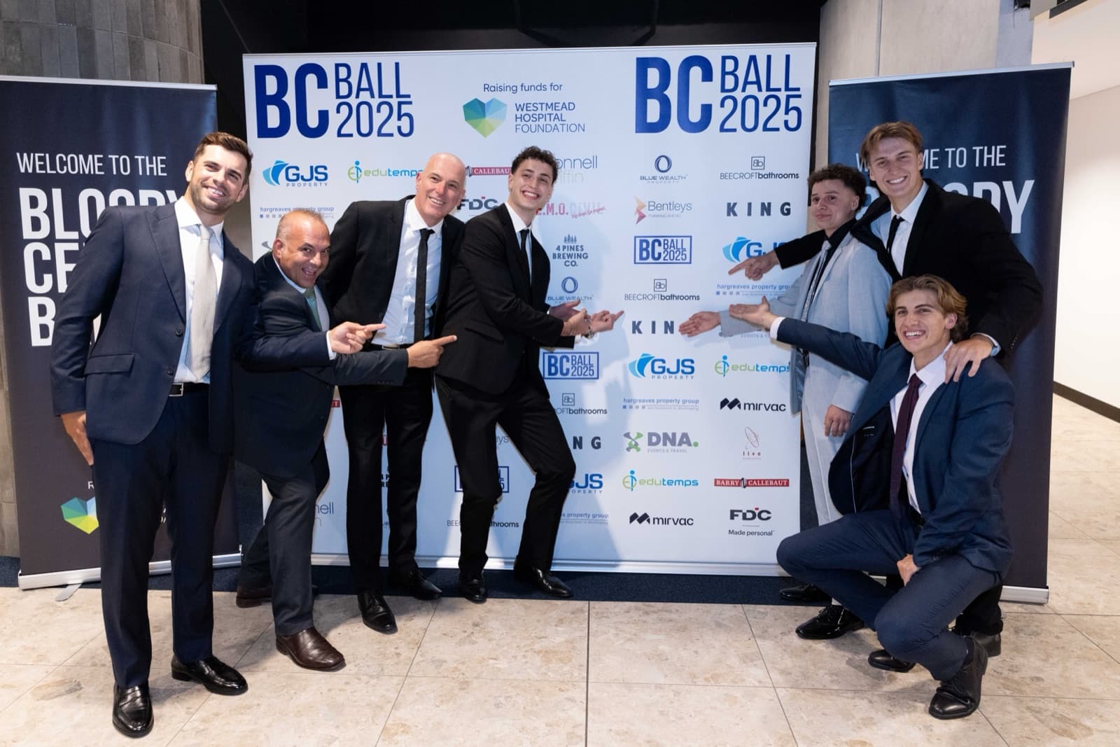 A group of sponsors playfully pointing at their company logos on the 2025 Bloody Cells Ball sponsor media wall