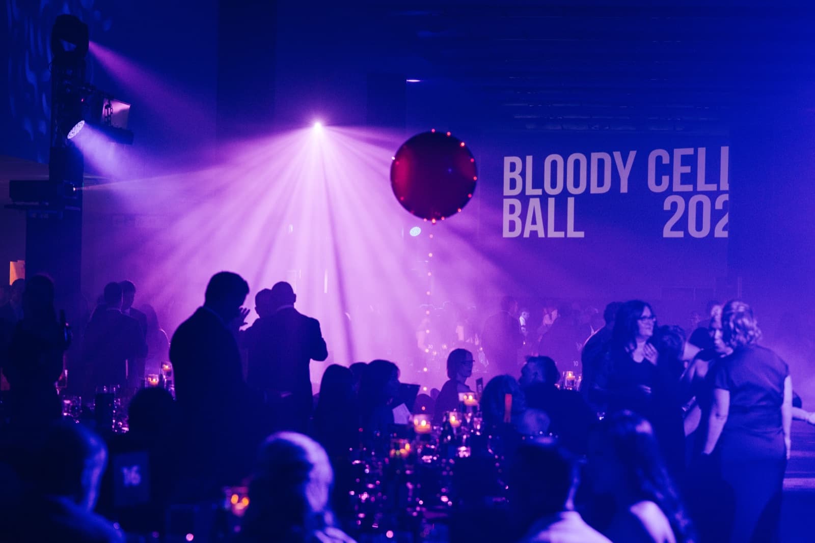 Bloody Cells Ball guests in a purple-lit ballroom with the event screen glowing in the background